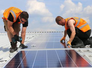 Two men in orange vests installing solar on the rooftop of a warehouse in the UK